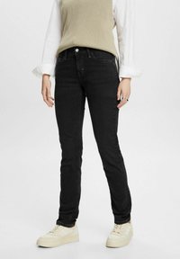 Jeans de mezclilla negra con corte ajustado, que cuentan con una bragueta de botón y cremallera estándar, combinados con una blusa de punto beige y zapatillas blancas.