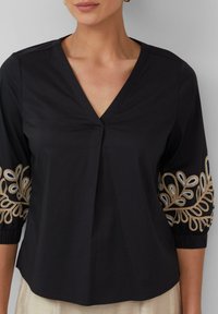Zwarte v-hals blouse met driekwartmouwen. Heeft complexe gouden en blauwe borduursels langs de mouwen. Glad materiaal met een relaxte pasvorm.