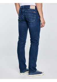 Férfi sötétkék egyenes szárú farmert és tengerészkék vászon tornacipőt visel, és egy egyszerű világosszürke háttér előtt áll.