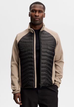 Volwassen man draagt een beige en zwarte jas met rits en doorgestikte voorkanten, staand tegen een effen lichtgrijze achtergrond.