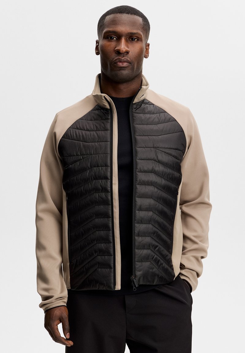 Voksent mand iført beige og sort lynlåstrøje med quiltede frontpaneler, stående mod en ensfarvet lysegrå baggrund.