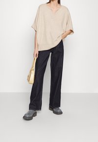 Beige übergroßer Strickpullover mit V-Ausschnitt, kombiniert mit dunkelblauen weiten Jeans. Graue Gummistiefel und eine strukturierte gelbe Handtasche runden das Outfit ab.