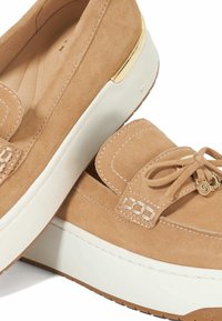 Beige suède loafers met een witte plateauzool, voorzien van een strikdetail en gouden accenten aan de zijkant. Gladde textuur met stiksels.