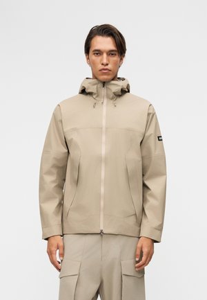 Muškarac odjeven u bež jaknu s kapuljačom zatvorenu patentnim zatvaračem i usklađene bež cargo hlače, stoji na jednostavnoj svijetloj pozadini.