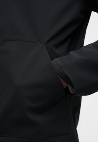 Αδιάβροχο μαύρο μπουφάν με απαλή υφή, διαθέτει τσέπη καγκουρό και ελαστικές μανσέτες για ασφαλή εφαρμογή.