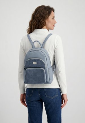 Kvinde med bølget brunt hår iført en hvid langærmet skjorte og blå jeans, der bærer en lyseblå læderrygsæk med forlommer.