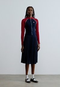 Tumesinise volditud seelikuga koos punast värvi pikkade varrukatega kitsas topp, millel ees lukk. Modell kannab musti kingi ja valgeid sokke. Siledapinnaline tekstuur.