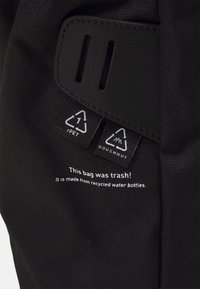 Musta reppu, joka on valmistettu kierrätysmateriaaleista. Reppu on kuvioitua kangasta, ja siinä on etikettejä sekä kestävyyttä osoittavaa painettua tekstiä. Yksityiskohdissa on tunnisteita.