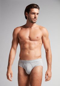 Graue Baumwollunterhose mit breitem Bündchen, das eine glatte Textur und doppelte Nähte entlang der Kanten aufweist. Das Modell steht mit einem neutralen Gesichtsausdruck.