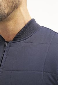 Mörk marinblå kviltad bomberjacka med ribbad krage, som har en slät yta och subtila sömdetaljer. Dragkedjestängning fram.