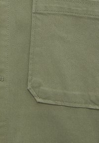 Tela verde oliva con un aspecto texturizado y tejido. Presenta un bolsillo con una solapa puntiaguda y costuras visibles a lo largo de los bordes.