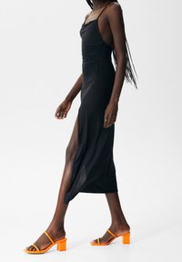 Svart midiklänning med sidoklyfta och rynkning, i kombination med orange blockklack-sandaler. Remmar och smal passform framhäver en elegant silhuett.