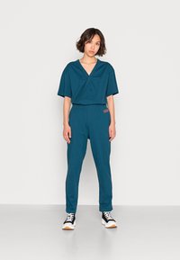 Jonge vrouw staat met een effen achtergrond in een teal wikkeltop en bijpassende broek met zwart-witte sneakers.