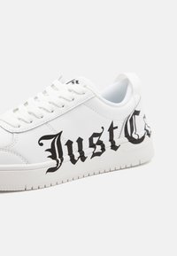 White sneaker with black graphic text "Just C" on the side, featuring a smooth, textured surface and flat laces.