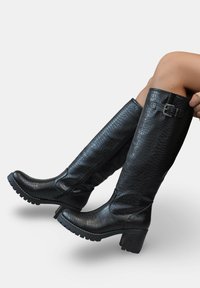 Bottes noires montantes avec un motif crocodile, fermeture éclair sur le côté, talon épais et détail de boucle sur le haut. La semelle en caoutchouc offre une bonne adhérence.
