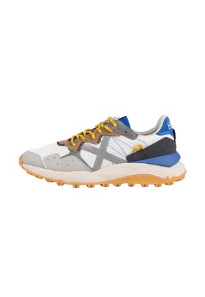 Zapatilla blanca y gris con cordones amarillos, detalle azul en el talón, suela marrón clara y una gran "X" gris en el lateral.