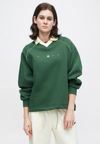 Pessoa com cabelo escuro na altura dos ombros usa um moletom verde da Nike sobre um colarinho amarelo pálido e calças brancas com cordões.