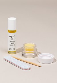 Nykkr Nighttime Nail & Cuticle Oil i en klar flaske, gul balsam i et rundtt glasskrukke, en blå neglefil og en trepinne på en nøytral bakgrunn.