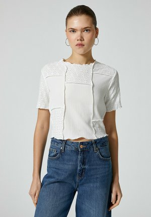 Hvid kortærmet bluse med en tekstureret overflade, der har hulsyninger og bølgede kanter, parret med høj talje denim jeans i blå.