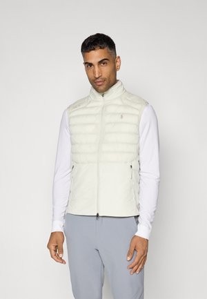 Gilet rembourré de couleur claire avec des coutures horizontales, un col montant et des poches zippées. Porté par-dessus une chemise blanche à manches longues.