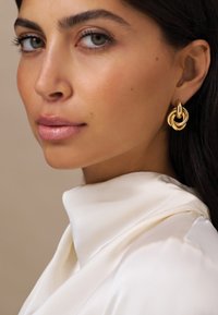 Goldene Creolenohrringe mit ineinandergeschachtelten kreisförmigen Designs, die eine glatte, glänzende Oberfläche aufweisen. Das Modell trägt ein weißes Satinhemd mit umgeschlagenem Kragen.