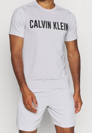 Homme portant un t-shirt blanc Calvin Klein et un short clair assorti, debout devant un fond uni.