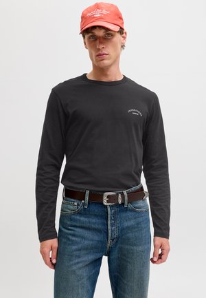 Joven con gorra coral, camiseta negra de manga larga con texto pequeño en el pecho, cinturón marrón oscuro y vaqueros azules sobre un fondo liso.