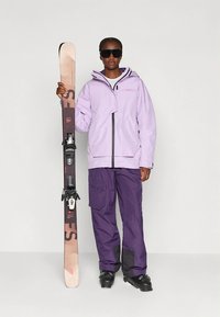 Hellviolette Skijacke mit schwarzen Akzenten, kombiniert mit dunkelvioletten Skihosen. Das Modell hält Skier mit einem schwarz-pinken Design.