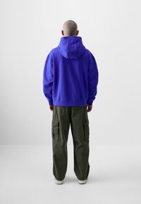 Felpa viola con cappuccio e vestibilità rilassata, abbinata a pantaloni cargo verde oliva con tasche laterali e coulisse in vita.