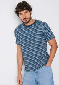 Camiseta a rayas en tonos azul marino y celeste con cuello redondo y mangas cortas, hecha de una tela suave. Se llevan vaqueros celestes.