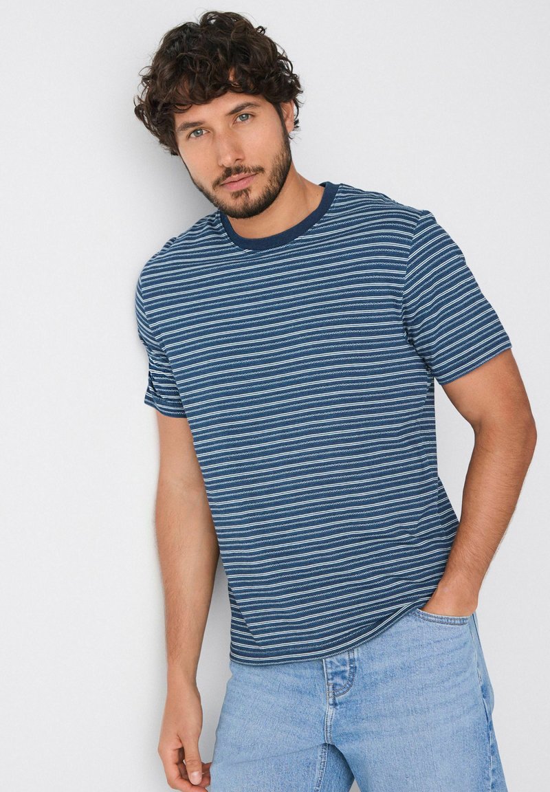 Camiseta a rayas en tonos azul marino y celeste con cuello redondo y mangas cortas, hecha de una tela suave. Se llevan vaqueros celestes.