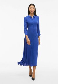 Modrá, dlhá šatka s dlhým rukávom vyrobená z hladkej tkaniny. Má predné gombíky, golier a splývavú sukňu. Nosí sa s čiernymi podpätkami.