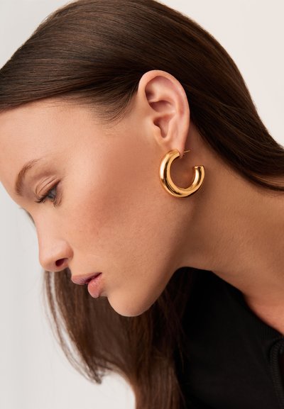 Boucles d'oreilles créoles dorées avec une surface lisse et polie, au design circulaire ouvert, portées à l'oreille, montrées de profil.