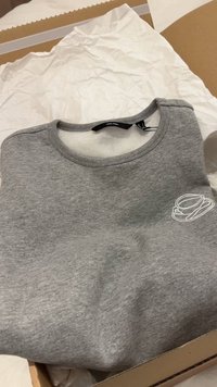 Πτυσσόμενο γκρι φούτερ με στρογγυλή λαιμόκοψη και μικρό λευκό σχέδιο στο στήθος, τοποθετημένο μέσα σε χάρτινο κουτί με λευκό χαρτί περιτυλίγματος.