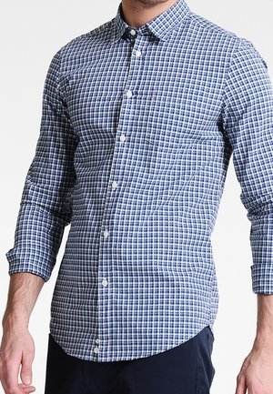 Homme portant une chemise à carreaux bleu et blanc à manches longues avec une poche poitrine et un pantalon bleu marine.