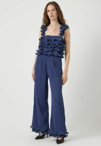 Combinaison bleu marine avec un haut texturé à volants et un pantalon à jambes larges. Présente un décolleté carré et des volants accentués sur les bretelles et les ourlets.