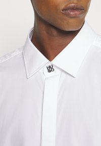 Gros plan sur une personne portant une chemise blanche à col avec une épingle en métal « HUGO » fixant le premier bouton.