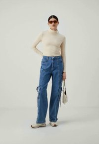 Donna che indossa un dolcevita beige, jeans cargo blu, sneakers bianche e occhiali da sole, mentre tiene una piccola borsa bianca su uno sfondo neutro.