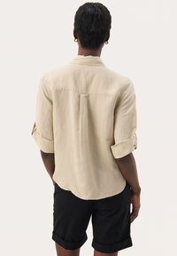 Camicia beige a maniche corte con risvolti, caratterizzata da una piega posteriore e una trama liscia, abbinata a pantaloni corti neri.