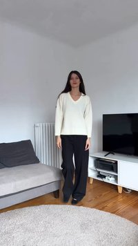 Κρεμ πουλόβερ με λαιμόκοψη V και ραμμένους ώμους, συνδυασμένο με μαύρο παντελόνι φαρδιάς γραμμής, τοποθετημένο σε ένα φωτεινό σαλόνι. Απαλές υφές και ουδέτεροι τόνοι.