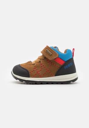 Pantofle din suede maro pentru copii cu capac de cauciuc negru la vârf, accente albastre și roșii, curea Velcro și detalii de broderie geometrică pe lateral.