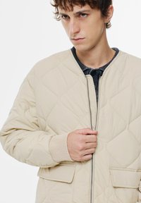 Beige quiltet bomberjakke med ribbet manchetter og en frontlynlås. Har to forlommer og et struktureret indre lag.