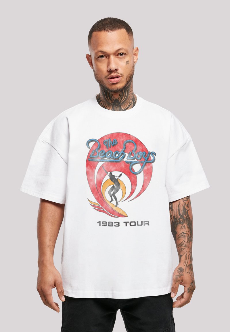 White oversized t-shirt featuring "The Beach Boys 1983 Tour" graphic with colorful surf design and retro font. Short sleeves and soft texture.