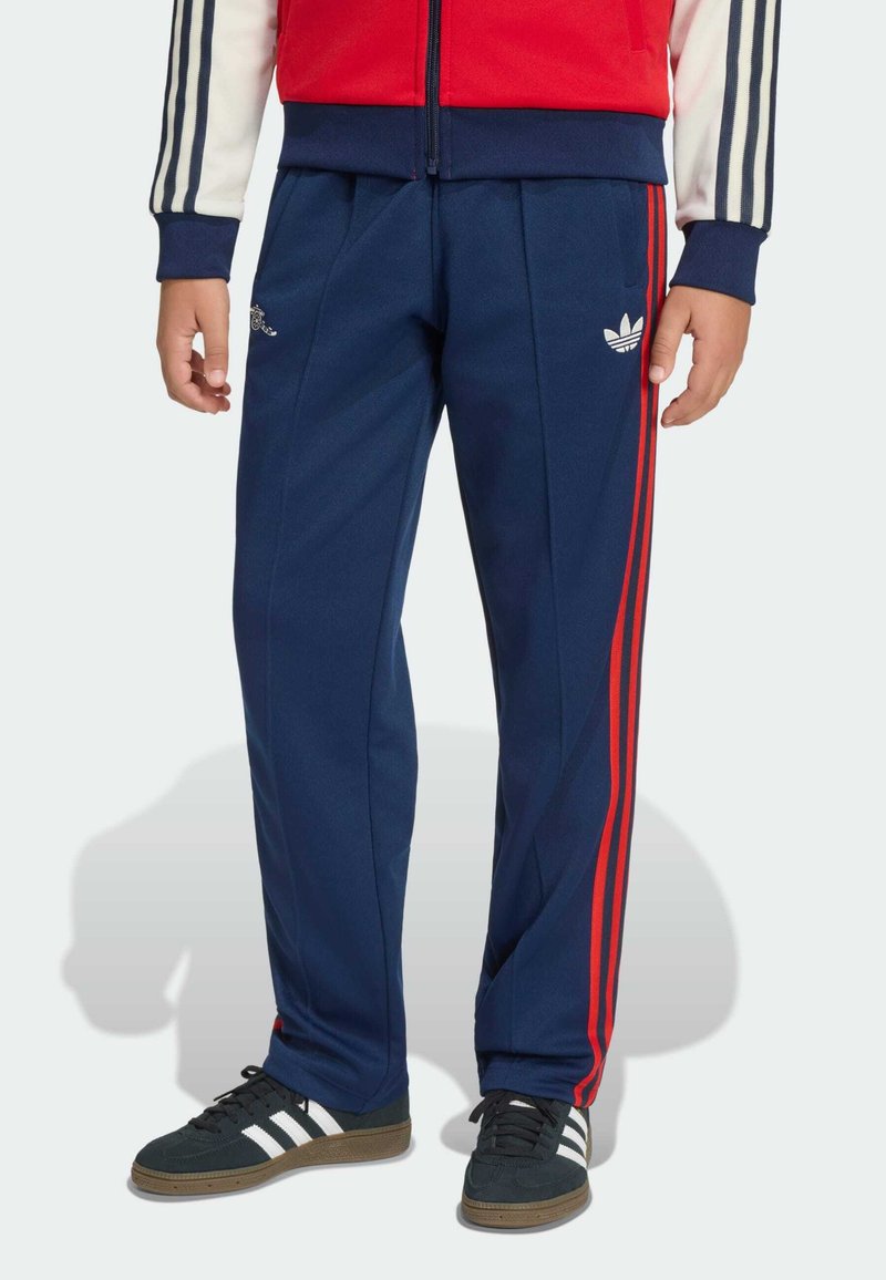 Παιδί που φοράει σκούρο μπλε αθλητικό παντελόνι με κόκκινες ρίγες, μαύρα αθλητικά παπούτσια με λευκές ρίγες και κόκκινο-λευκό μπουφάν με σκούρες μπλε μανσέτες.