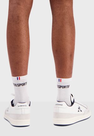 White leather sneakers with grey rubber sole, navy accents, and logo on the heel, paired with white socks featuring a blue-red flag detail.