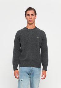 En ung man med kort mörkt hår bär en mörkgrå tröja med rundad hals och en liten grön logotyp samt ljusblå slitna jeans, står mot en vit bakgrund.