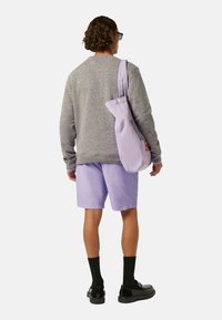 Grå strikket sweatshirt, lyse lilla shorts, sorte sokker, sorte sko, bærer en lila tote bag med en lille logo detalje.