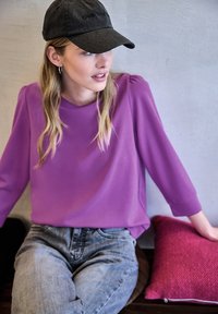 Maglia lunga viola con scollatura tonda, abbinata a jeans chiari strappati. Cappellino da baseball scuro e un cuscino rosa testurizzato sullo sfondo.