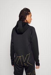 Zwarte hoodie met gele geometrische logo-accenten, met een gladde textuur en een getailleerd ontwerp, getoond vanuit de achterkant.