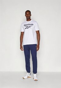 Biała koszulka Reebok z krótkim rękawem i granatowym logo, w połączeniu z granatowymi dresami i białymi trampkami z gumowymi podeszwami, stojąca na neutralnym tle.